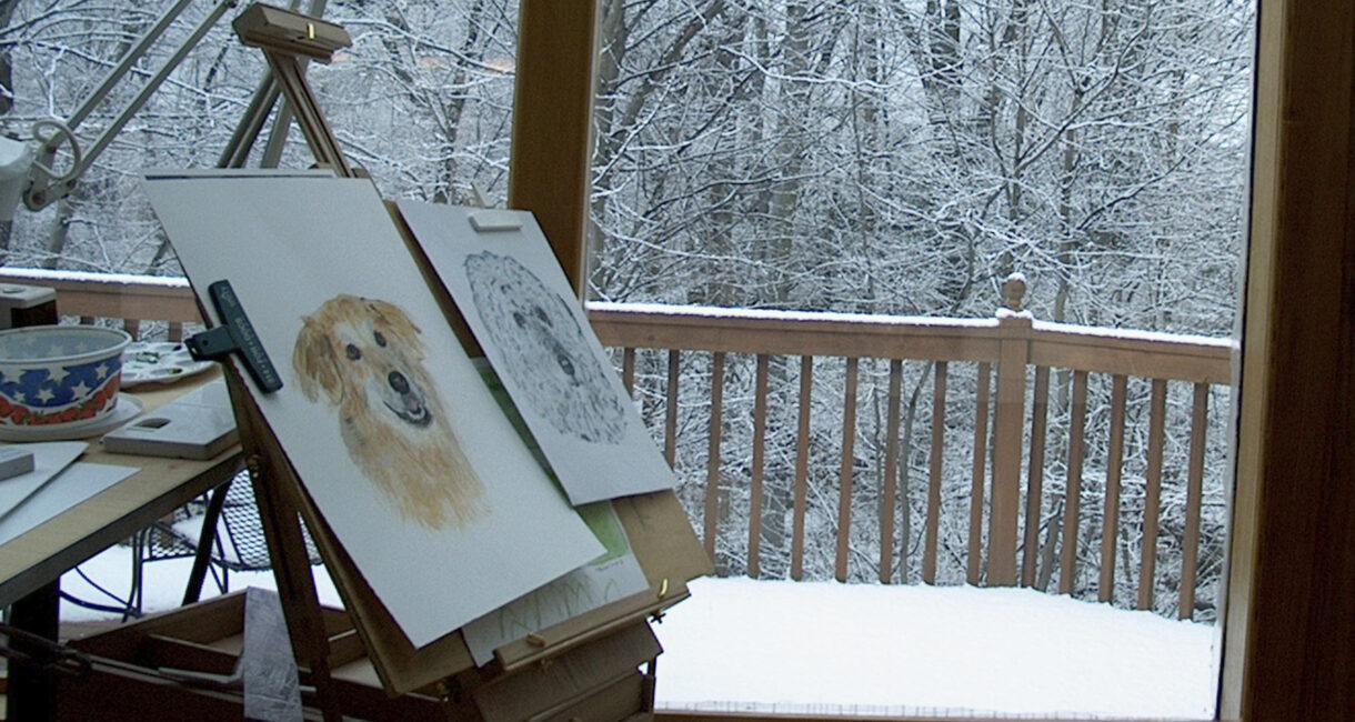 Behind the story of Promises Kept shows a drawing board in front of a window with a snowy landscape outside, featuring sketches of various dogs , creating a cozy and artistic scene with wintery charm