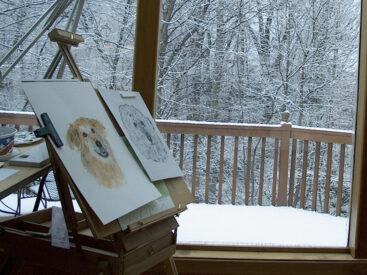 Behind the story of Promises Kept shows a drawing board in front of a window with a snowy landscape outside, featuring sketches of various dogs , creating a cozy and artistic scene with wintery charm